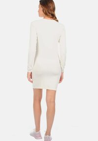 Robe crème à manches longues avec une silhouette ajustée et une texture côtelée. L'ourlet arrive au milieu de la cuisse. Associée à des baskets rose clair.