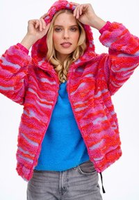 Miękka, różowa i fioletowa kurtka na zamek błyskawiczny z kapturem, z falistym wzorem i delikatną fakturą, noszona na niebieskim swetrze.