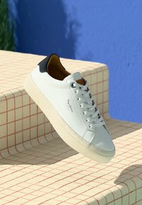 White leather sneaker with black heel tab, white laces, and beige sole, resting on beige tiled steps with red grid lines and blue wall background.