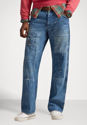 Kék farmernadrág több folttal, laza szabással és látható festékfröccsenésekkel. Gombos és cipzáras záródással, valamint oldalzsebekkel rendelkezik.