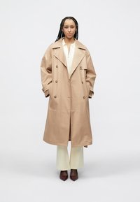 Μπεζ παλτό τύπου trench με φαρδιές πέτσες γιακά, διπλό κούμπωμα και μεγάλα κουμπιά, συνδυασμένο με λευκή μπλούζα και εκρού παντελόνι.