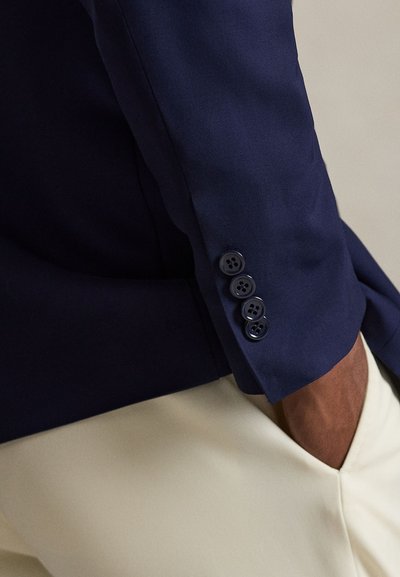 Tengerészkék blézerujj négy sötét gombbal, bézs nadrágon pihenve, a kéz részben a zsebben, semleges háttér előtt.