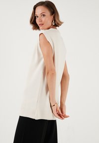 Cream-colored sleeveless top with a ribbed hem and side slits. Smooth texture, relaxed fit, paired with black trousers.