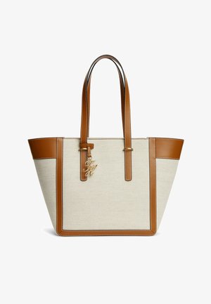 Beige Canvas-Tragetasche mit braunen Lederbesätzen, doppelten Schulterriemen und einem goldfarbenen Anhänger mit Initialen, der nahe der Oberseite hängt.