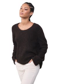 Naine patsidega juustega, kandes tumepruuni karvast kampsunit ja valgeid pükse, käed taskutes, vaadates veidi kõrvale valgel taustal.