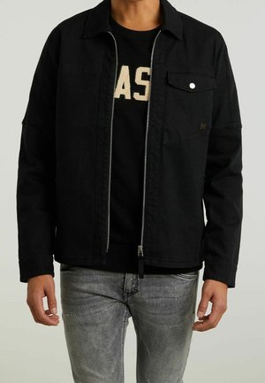 Homme portant une veste noire zippée sur un t-shirt noir avec des lettres beiges, associée à un jean gris clair usé, debout devant un fond uni.
