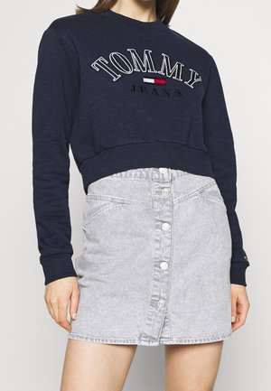 Vrouw draagt een navy Tommy Jeans cropped sweatshirt en een lichtgrijze high-waisted denim rok met knopen voor, staand tegen een effen achtergrond.