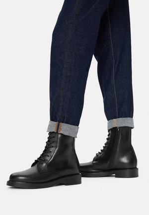 Schwarze Leder-Schnürstiefeletten mit seitlichem Reißverschluss, getragen zu dunkelblauen, umgeschlagenen Jeans vor einfachem Hintergrund.