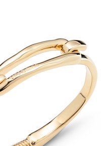 Bracelet en métal doré avec un design lisse et incurvé. Il présente un fermoir à extrémité arrondie avec un logo gravé. Apparence minimaliste et élégante.