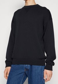 Schwarzes Langarm-Sweatshirt mit gerippter Textur und Rundhalsausschnitt. Verfügt über einen dezenten Akzent am Bündchen und wird mit blauen Jeans kombiniert.