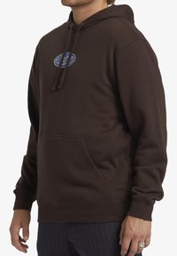 Sudadera marrón hecha de un tejido suave, con un bolsillo frontal, capucha con cordones y un logo colorido en el pecho. Corte holgado y puños de canalé.