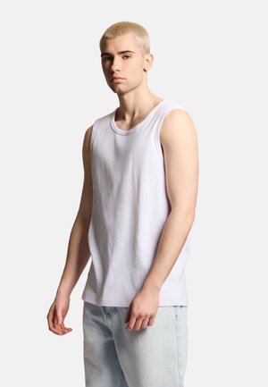 Młody mężczyzna z krótkimi blond włosami, ubrany w białą koszulkę bez rękawów i jasnoniebieskie dżinsy, stoi na prostym jasnym tle.