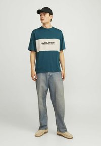 Kortærmet teal t-shirt med creme vandret stribe og sort tekst. Sammen med lyseblå løstsiddende jeans og beige sko.