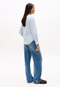 Hosszú sötét hajú nő világoskék, gombos ingben, bő szárú kék farmerben és sötét mokaszinban, oldalra fordulva, fehér háttér előtt áll.