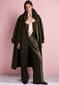 Olive green wool coat with large lapels, button details, tailored green suit, and light pink bow tie blouse. Glossy black shoes.