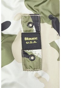 Tela de camuflaje con varios patrones en verde, negro y beige. Presenta una etiqueta negra que dice "Blauer: U.S.A." con texto amarillo y dos ojetes metálicos.