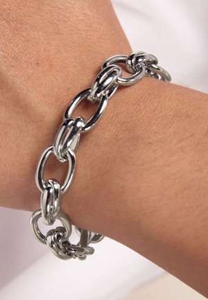 Bracciale in metallo d'argento con maglie ovali e rotonde interconnesse, texture liscia e finitura lucida, indossato al polso.