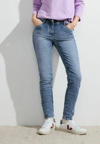 Jean skinny bleu clair avec un délavage, comportant cinq poches et une taille mi-haute, associés à des baskets blanches avec des accents rouges.