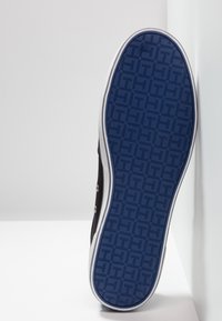 Sohle eines schwarzen Sneakers mit blauem Muster, weiße Zwischensohle, vertikal an einer weißen Ecke Wand und Boden lehnen.