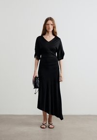 Γυναίκα που φοράει μαύρο φόρεμα με ντεκολτέ σε σχήμα V, μανίκια μέχρι τον αγκώνα και ασύμμετρο τελείωμα, κρατώντας μια μικρή μαύρη τσάντα με κορδόνι, στέκεται σε απλό δάπεδο.