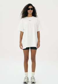 Witte oversized t-shirt met korte mouwen en logo in het midden, gecombineerd met zwarte shorts, witte sokken en veelkleurige sneakers.