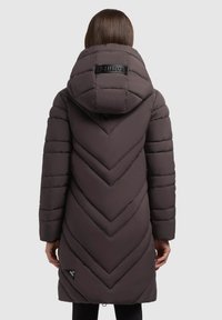 Donna che indossa un lungo cappotto invernale imbottito scuro con un grande cappuccio e trapuntatura a zigzag, vista di spalle su uno sfondo neutro.
