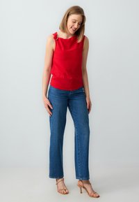 Top rosso senza maniche con un dettaglio decorativo a nodo sulla spalla, abbinato a jeans a gamba larga blu chiaro e sandali nude con fascia.