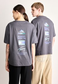 T-Shirts cinzentas de algodão com corte descontraído, apresentando gráficos de logotipo em azul, branco e verde nas costas, combinadas com calças bege claro.