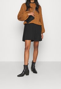 Maglione marrone a maglia, vestito nero svasato e stivali neri alla caviglia con tacco a blocco. Piccola borsa nera con tracolla a catena.