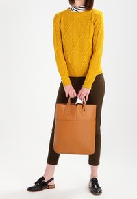 Tan läder toteväska med två handtag, rektangulär form och sömdetaljer. Modellen bär en senapsgul tröja och olivgröna byxor, svarta skor.