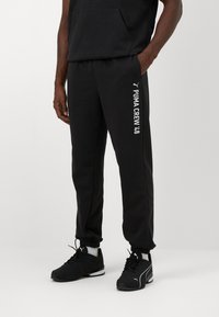 Man in zwarte Puma sweatpants met witte tekst "PUMA CREW '48", gecombineerd met zwarte Puma sneakers en witte sokken, staande met handen in de zakken.