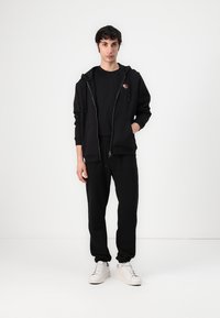 Sudadera negra con cremallera y bordado de rosa en el pecho, combinada con una camiseta negra y pantalones de chándal negros a juego. Zapatillas blancas.