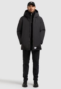 Fekete, párnázott kabát kapucnival, elülső cipzárral és oldalsó zsebekkel, fekete farmernadrággal és fekete csizmával párosítva. Egyszerű, funkcionális dizájn.