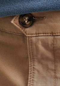 Pantaloni di stoffa marrone con una trama liscia, caratterizzati da un bottone prominente e cuciture a vista. Sopra, è visibile una camicia azzurra con texture.