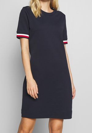 Mujer con un vestido azul marino hasta la rodilla y mangas cortas con franjas rojas, blancas y azules en los puños, sobre un fondo liso.