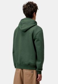 Sweat à capuche vert en coton doux avec une capuche spacieuse, des poignets et une taille côtelés. Porté avec un pantalon couleur beige.