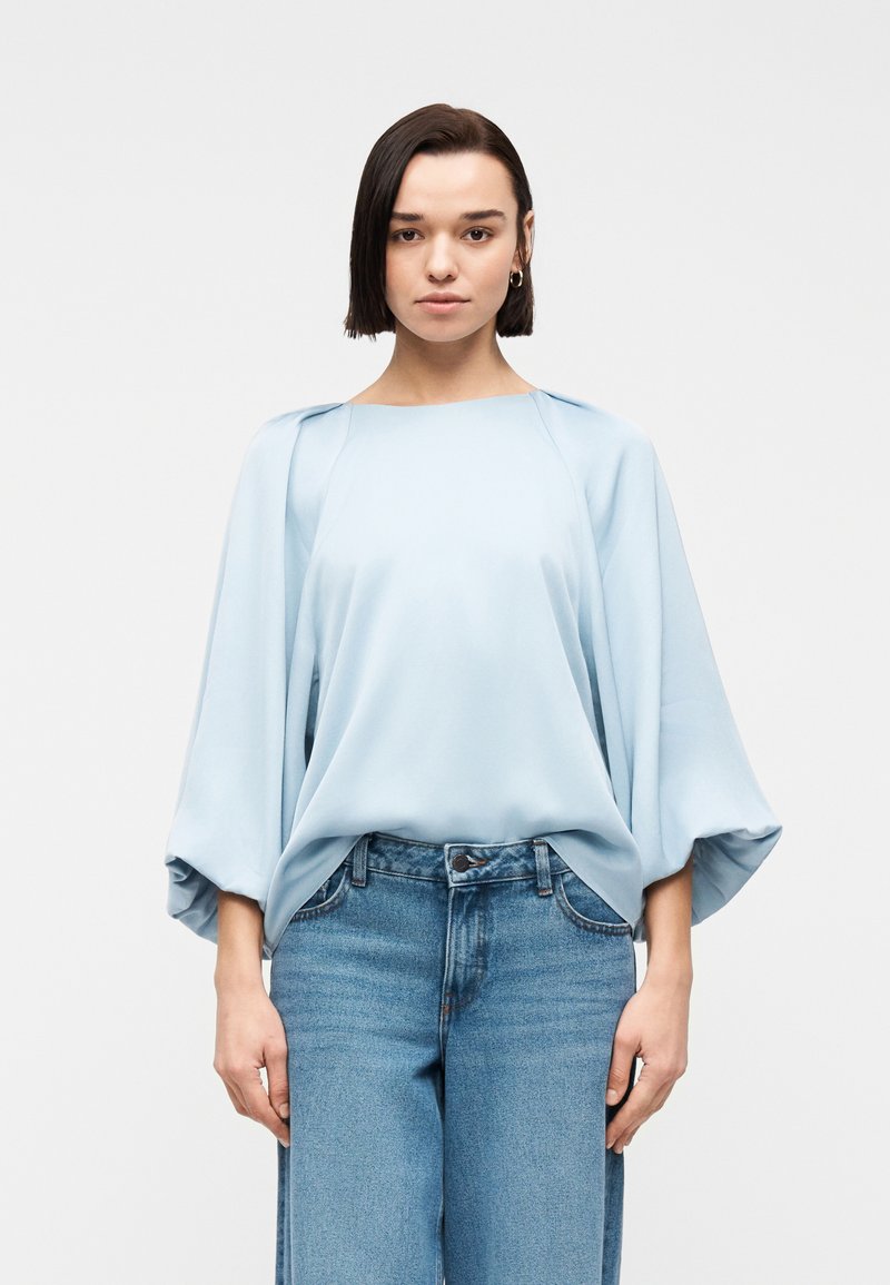 Γυναίκα με κοντά σκούρα μαλλιά, που φοράει ανοιχτό μπλε φαρδιά μπλούζα με φαρδιά μανίκια και μπλε τζιν παντελόνι, στέκεται μπροστά σε λευκό φόντο.