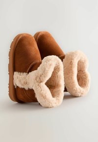Brune ruskinds slip-on støvler med en fluffy beige foring, der har en rund åbning og en struktureret gummisål for greb.