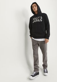 Crna kapuljača s grafičkim logotipom "JACK & JONES", sivi kadifasti hlače i crne tenisice s bijelim pertlama. Model stoji ispred jednobojne pozadine.