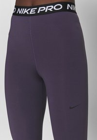 Fioletowe legginsy Nike Pro z czarnym elastycznym pasem, na którym znajduje się napis "NIKE PRO" w białym kolorze, gładka tekstura oraz subtelne logo Nike na nodze.