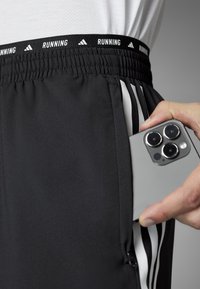 Schwarze Sportshorts aus strukturiertem Material mit einem elastischen Taillenbund und dem Aufdruck "RUNNING", weißen Akzenten und einer Seitentasche, die ein Smartphone hält.