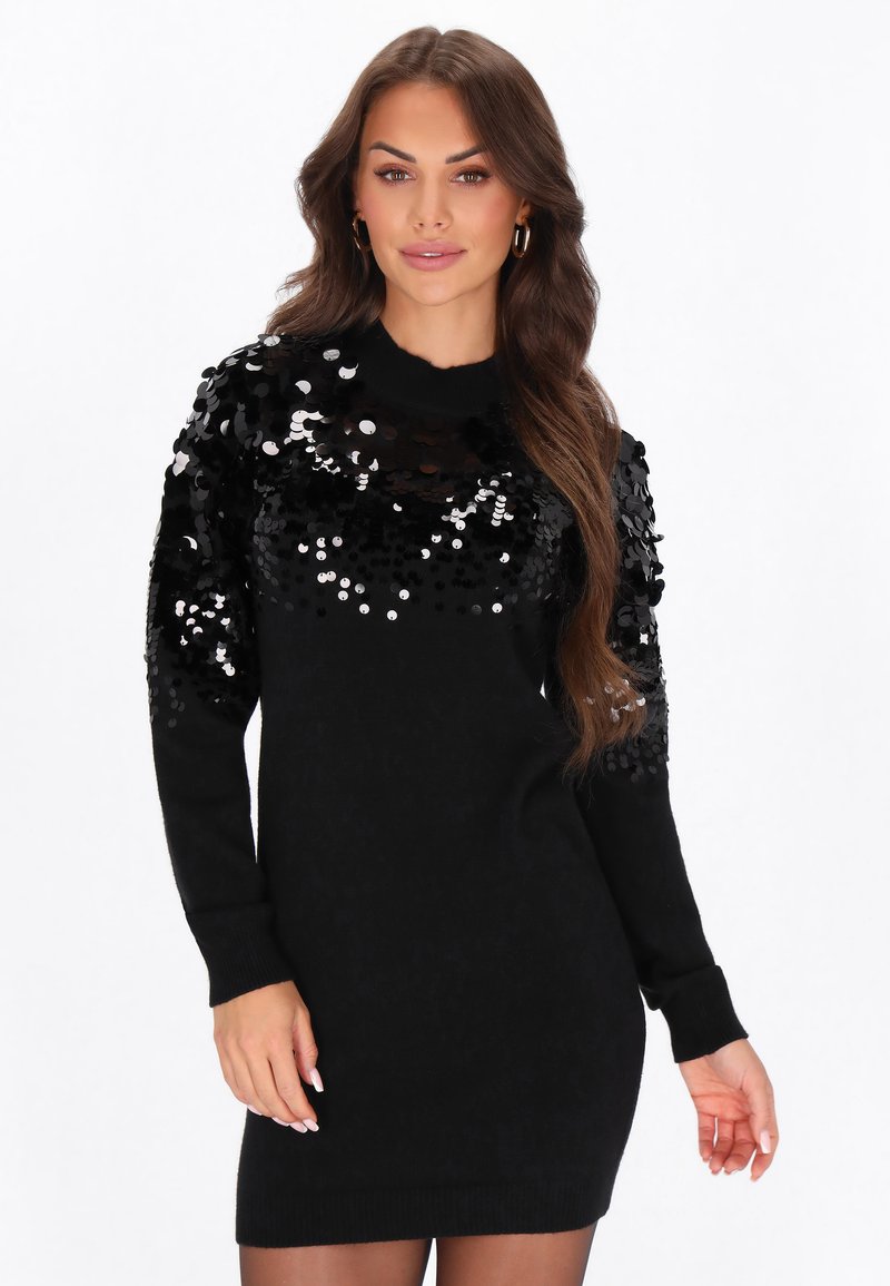 Donna che indossa un vestito maglione nero a maniche lunghe con paillettes nere lucide sul petto e sulle maniche, in piedi davanti a uno sfondo bianco.