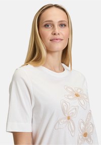 T-shirt en coton blanc à manches courtes, avec des motifs floraux brodés en beige clair et des accents dorés à l'avant.