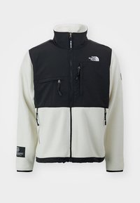 Fleecejacke mit weißem Körper und schwarzem Oberteil, ausgestattet mit Reißverschlusstaschen, einem hohen Kragen und einem Logo auf der Brust. Strukturierter Stoff.