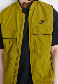 Man som bär en olivgrön Nike-väst med flera dragkedjefickor över en svart kortärmad skjorta, visad från axlarna till midjan.