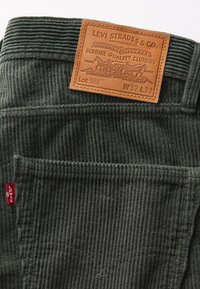 Mörkgröna kordbyxor har en strukturerad yta, läderpatch med varumärkesdetaljer och röd logotag på sidan.