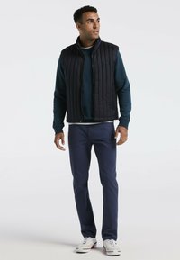 Schwarze gesteppte Weste über einem marineblauen Pullover, kombiniert mit marineblauen Hosen und weißen Sneakern. Einfaches Design mit funktionalen Taschen und glatter Textur.
