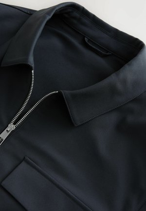 Schwarzes Poloshirt mit Reißverschluss und Stehkragen, aus glattem Stoff mit dezentem Glanz. Verfügt über eine Fronttasche und metallische Reißverschlussdetails.