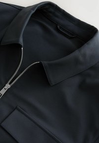 Black zippered polo shirt with a stand collar, smooth fabric, and subtle sheen. Features a front pocket and metallic zipper detail.