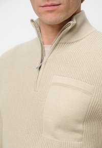 Beige Strickpullover mit einem Reißverschlusskragen und einer Vordertasche, ausgestattet mit einem strukturierten Muster und gerippten Details entlang der Kanten.